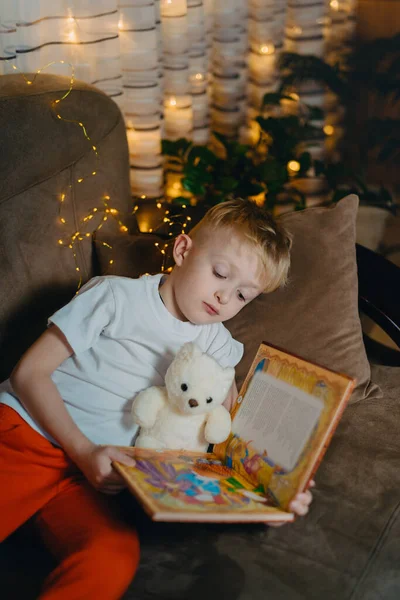 3-4 years old boy is reading a book sitting on a sofa with teddy bear. Home reading of fairy tales. Cute little boy is reading book while sitting on a sofa. Child reads a book to his best bear friend. — Stockfoto