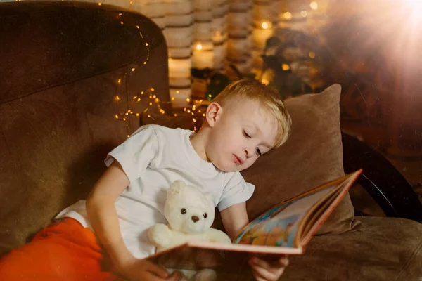 3-4 years old boy is reading a book sitting on a sofa with teddy bear. Home reading of fairy tales. Cute little boy is reading book while sitting on a sofa. Child reads a book to his best bear friend. — Stock Photo, Image