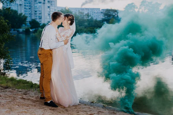 Bride and groom with the blue and yellow colored smoke bombs in the summer park. The bride and groom with the colored smoke at the background of summer forest. Wedding couple runs color smoke in the — Fotografia de Stock