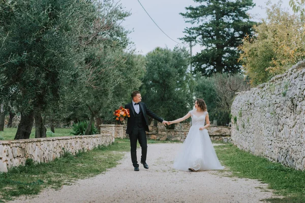 Glückliches, stylisch lächelndes Paar bei einem Spaziergang in der Toskana, Italien an ihrem Hochzeitstag. Braut und Bräutigam gehen an den Händen die Straße hinunter. Ein stilvolles junges Paar geht spazieren. Mann und Frau kommunizieren gut — Stockfoto