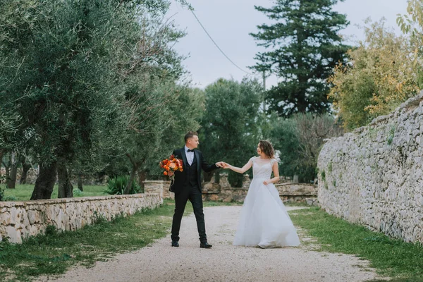 Glückliches, stylisch lächelndes Paar bei einem Spaziergang in der Toskana, Italien an ihrem Hochzeitstag. Braut und Bräutigam gehen an den Händen die Straße hinunter. Ein stilvolles junges Paar geht spazieren. Mann und Frau kommunizieren gut — Stockfoto