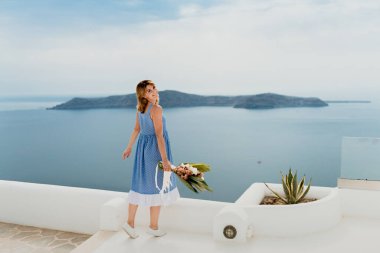 Yunanistan 'ın eski bir Avrupa kenti olan Santorini adasının caddesi boyunca çiçek buketiyle yürüyen güzel bir kız. Sokakta yürüyen bir turist kızın portresi. Aşk hikayesi yazı.