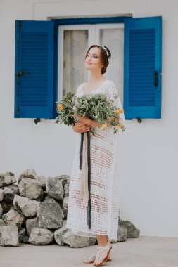 Beyaz duvarın yanında mavi pencereli güzel beyaz bir gelin poz veriyor. Çiçek buketiyle genç ve çekici gelin