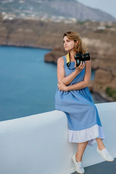 Vrouw in een blauwe jurk zittend op de rotsen op het strand en foto 's nemend met een SLR camera. Zee en bergen op de achtergrond — Stockfoto