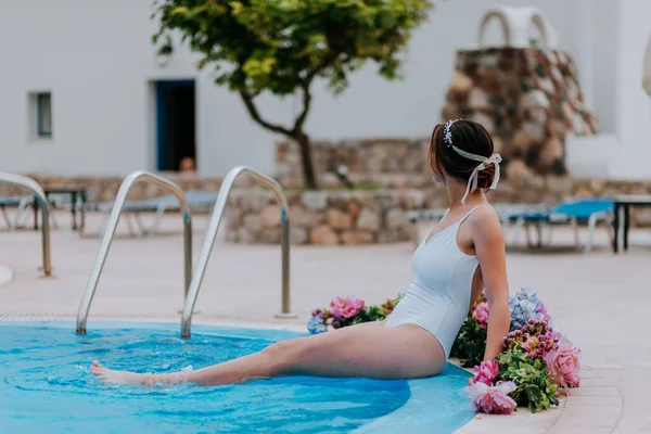 Hermosa novia en un hermoso traje de baño blanco y guirnaldas de flores, de pie en la piscina con flores en agua turquesa, hermosa chica en la piscina de agua en el día soleado — Foto de Stock
