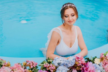 Güzel beyaz mayo ve çiçek çelenkleri içinde güzel bir gelin, turkuaz suda çiçekle yüzme havuzunda duruyor, güneşli bir günde su havuzunda güzel bir kız.