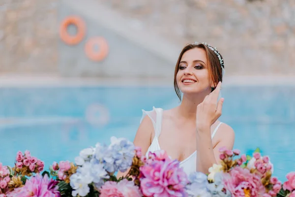 Hermosa novia en un hermoso traje de baño blanco y guirnaldas de flores, de pie en la piscina con flores en agua turquesa, hermosa chica en la piscina de agua en el día soleado — Foto de Stock
