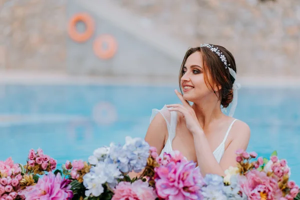 Hermosa novia en un hermoso traje de baño blanco y guirnaldas de flores, de pie en la piscina con flores en agua turquesa, hermosa chica en la piscina de agua en el día soleado — Foto de Stock