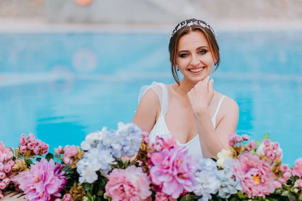Hermosa novia en un hermoso traje de baño blanco y guirnaldas de flores, de pie en la piscina con flores en agua turquesa, hermosa chica en la piscina de agua en el día soleado —  Fotos de Stock