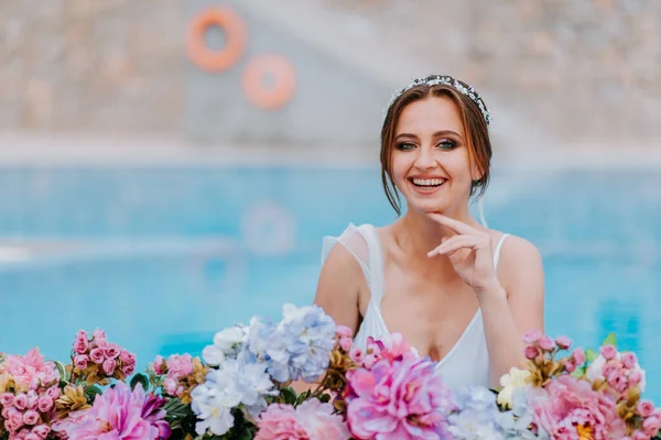 Hermosa novia en un hermoso traje de baño blanco y guirnaldas de flores, de pie en la piscina con flores en agua turquesa, hermosa chica en la piscina de agua en el día soleado —  Fotos de Stock