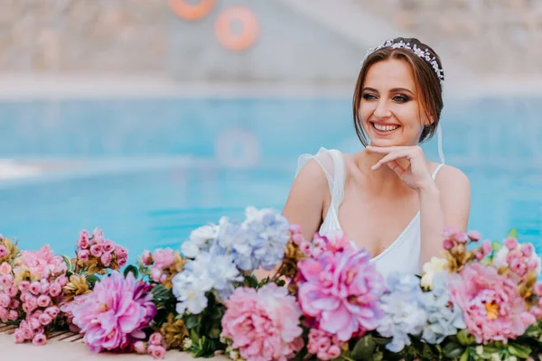 Pengantin cantik dengan baju renang putih yang indah dan karangan bunga, berdiri di kolam renang dengan bunga di atas air pirus, gadis cantik di kolam air di hari yang cerah — Stok Foto
