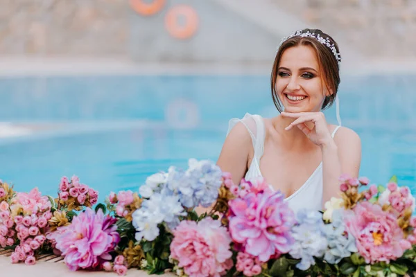 Hermosa novia en un hermoso traje de baño blanco y guirnaldas de flores, de pie en la piscina con flores en agua turquesa, hermosa chica en la piscina de agua en el día soleado —  Fotos de Stock