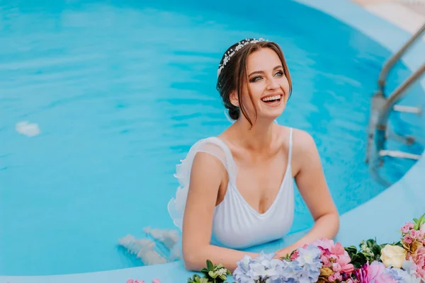 Belle mariée dans un beau maillot de bain blanc et des guirlandes de fleurs, debout dans la piscine avec des fleurs sur l'eau turquoise, belle fille dans la piscine d'eau dans la journée ensoleillée — Photo