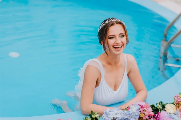 Belle mariée dans un beau maillot de bain blanc et des guirlandes de fleurs, debout dans la piscine avec des fleurs sur l'eau turquoise, belle fille dans la piscine d'eau dans la journée ensoleillée — Photo