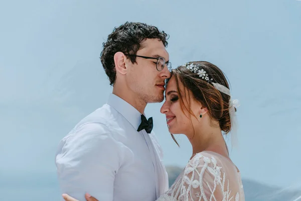 Красивые невеста и жених обнимаются и целуются под завесой невесты в день их летней свадьбы на греческом острове Санторини — стоковое фото
