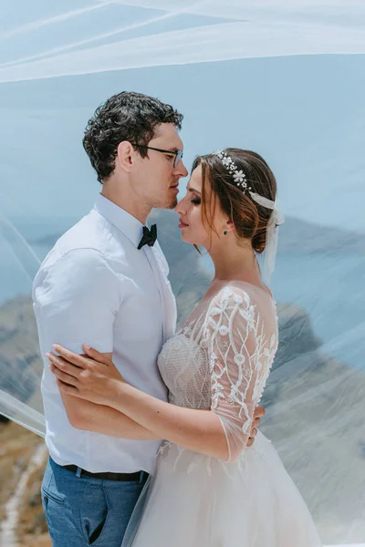 Bela noiva e noivo abraçando e beijando sob o véu nupcial em seu dia de casamento de verão na ilha grega Santorini — Fotografia de Stock