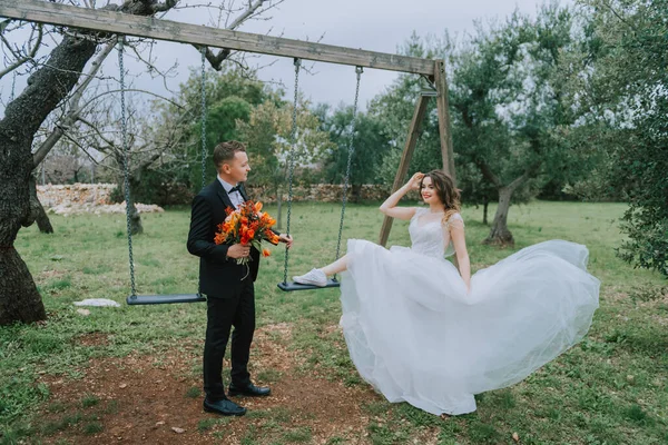 Felice elegante coppia sorridente passeggiando in Toscana, Italia il giorno del loro matrimonio. NEWLYWEDS con l'altalena nel parco. Gli sposi camminano per strada per le mani. Una giovane coppia alla moda cammina — Foto Stock