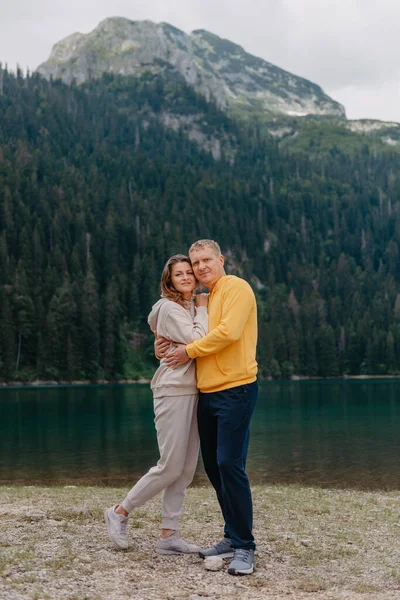 Ρομαντικό ζευγάρι στην αγάπη στέκεται στην πετρώδη ακτή της λίμνης, Γραφική θέα στο βουνό. Η νύφη και ο γαμπρός. Όμορφο καυκάσιο ζευγάρι αγκαλιάζεται από τη λίμνη την ηλιόλουστη μέρα του καλοκαιριού. Γαμήλιο ταξίδι — Φωτογραφία Αρχείου