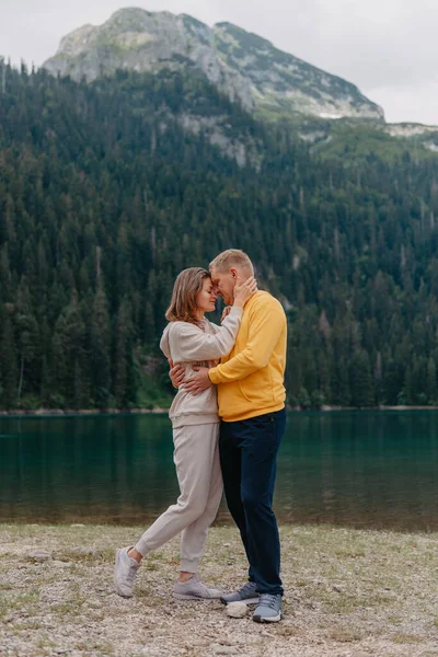 Ρομαντικό ζευγάρι στην αγάπη στέκεται στην πετρώδη ακτή της λίμνης, Γραφική θέα στο βουνό. Η νύφη και ο γαμπρός. Όμορφο καυκάσιο ζευγάρι αγκαλιάζεται από τη λίμνη την ηλιόλουστη μέρα του καλοκαιριού. Γαμήλιο ταξίδι — Φωτογραφία Αρχείου