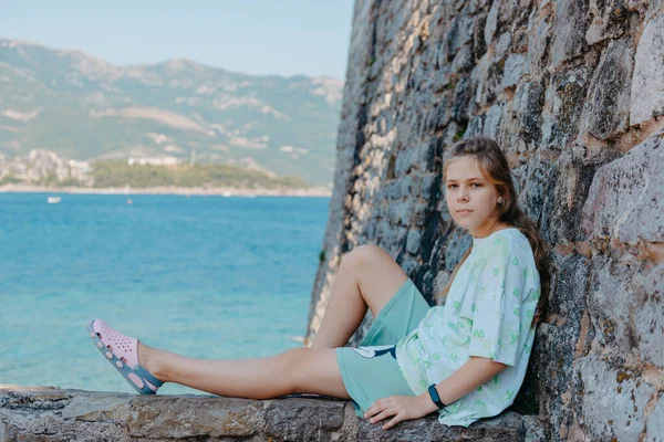 Mooi meisje zit op een stenen muur, op de achtergrond is de blauwe zee, Budva, Montenegro. — Stockfoto