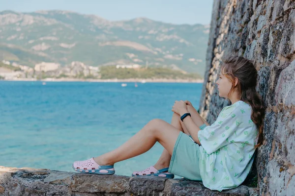 Hermosa chica sentada en una pared de piedra, en el fondo es el mar azul, Budva, Montenegro. — Foto de Stock