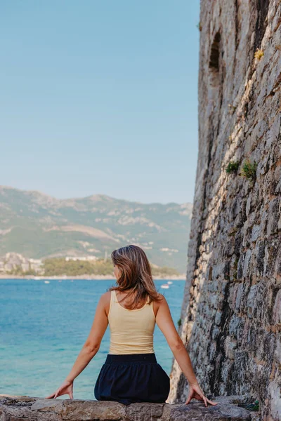 Hermosa chica sentada en una pared de piedra, en el fondo es el mar azul, Budva, Montenegro. — Foto de Stock