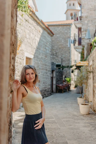 Meisje wandelend door de oude smalle straat op een mooie zomerdag in MEDITERRANEAN MEDIEVAL CITY, oude stadswijk BUDVA, MONTENEGRO. Jonge mooie vrolijke vrouw wandelen op de oude straat op — Stockfoto