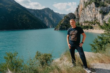 Güneşli ve huzurlu bir günün alacakaranlıkta gölde yakışıklı bir genç adam, Karadağ Alpleri 'ndeki Piva Gölü kıyısının yanındaki kayanın yanında duran bir adam. Dağlar. Yaz yürüyüşü. Yaz Harikalar Diyarı