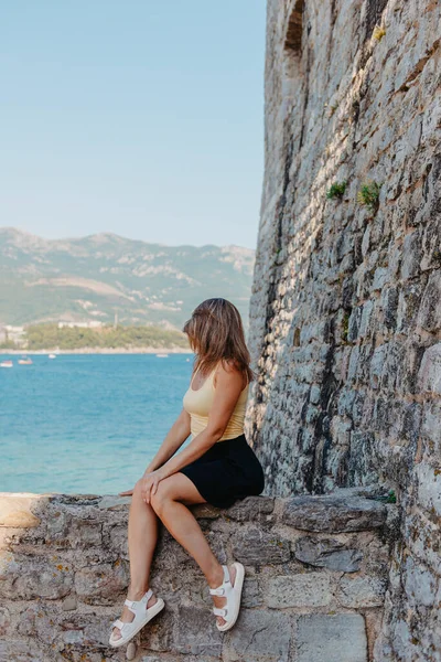 Vacker flicka sitter på en stenmur, i bakgrunden är det blå havet, Budva, Montenegro. — Stockfoto