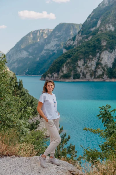 Chica turística se encuentra en la orilla de un lago en las montañas. hermoso paisaje, Lago Piva en Montenegro.. Mujer está de pie en la costa del lago Piva al atardecer en verano. Paisaje con chica, famoso — Foto de Stock