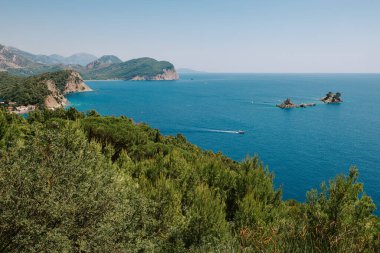 Denizden güneşli bir günde kayaların üzerinde resim gibi bir manzara. Budva Riviera, Karadağ. Karadağ 'ın Budva adası Sveti Nikola' nın hava manzarası. Hawaii plajı, şemsiyeler, banyolar ve kristal berrak sular