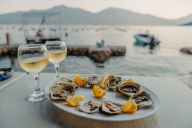 Tabakta taze yakalanmış istiridye ve asma bardakları. Karadağ 'daki istiridye çiftliği yakınlarındaki Kotor Körfezi kıyısında bir restoran. Deniz ürünleri. İstiridye çiftliği ve dağlarıyla güzel bir deniz manzarası, Adriyatik