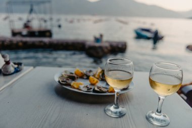 Tabakta taze yakalanmış istiridye ve asma bardakları. Karadağ 'daki istiridye çiftliği yakınlarındaki Kotor Körfezi kıyısında bir restoran. Deniz ürünleri. İstiridye çiftliği ve dağlarıyla güzel bir deniz manzarası, Adriyatik