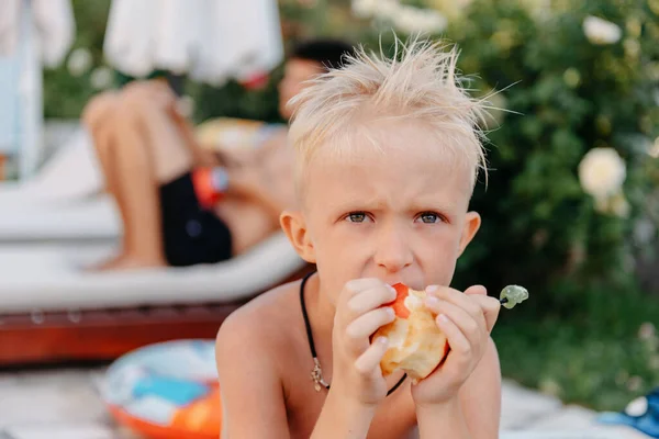Riéndose lindo niño ucraniano en el camping sentado y comer fruta de manzana. Retrato de niño sonriente feliz en el fondo de la naturaleza. Chico gracioso. Niño comiendo fruta de manzana verano al aire libre — Foto de Stock