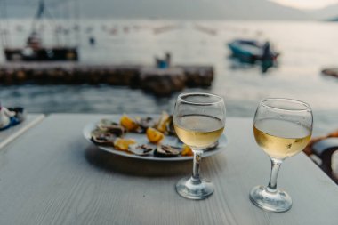Tabakta taze yakalanmış istiridye ve asma bardakları. Karadağ 'daki istiridye çiftliği yakınlarındaki Kotor Körfezi kıyısında bir restoran. Deniz ürünleri. İstiridye çiftliği ve dağlarıyla güzel bir deniz manzarası, Adriyatik