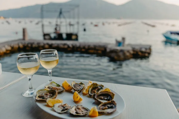 Ostras recién capturadas en un plato y vasos de vid. Restaurante en las orillas de la bahía de Kotor cerca de la granja de ostras, Montenegro. Mariscos. Hermoso paisaje marino con granja de ostras y montañas, Adriático — Foto de Stock