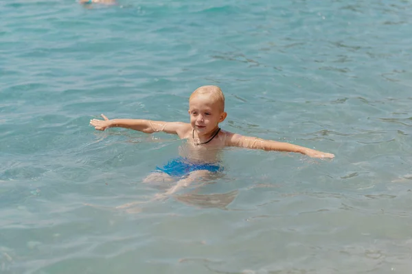 Niño aprendiendo a nadar en el mar abierto del resort tropical. Los niños aprenden a nadar. Ejercicio y entrenamiento para niños pequeños. Niño pequeño con tablero de flotador de colores en el club deportivo. Nadar bebé o niño pequeño — Foto de Stock