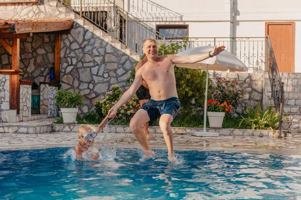 Excited boy in googles jumping in water from shoulders of his father standing in swimming pool