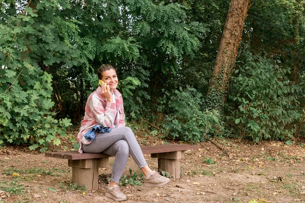快乐的女孩坐在长椅上 在街上用智能手机 美丽的女孩坐在长椅上 时尚的生活方式和你的手机一起在社交网络上留言 在公园里阳光灿烂的一天 — 图库照片
