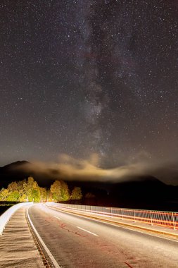 milky way in night sky above street with light trails in alps