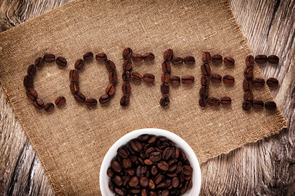 Kahve çekirdekleri bir çuval bezi üzerinde kelime "kahve" olarak. ahşap arka plan — Stok fotoğraf