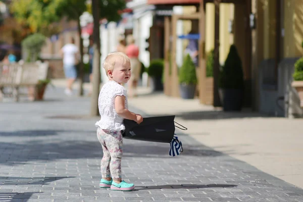 Linda niña de pie o caminando en el medio de la calle en la aldea de salida durante las ventas con bolsa de compras negro en sus manos, multitud de personas en el fondo —  Fotos de Stock