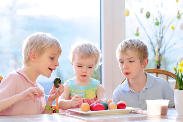 Groep van schattige kinderen van één familie, twee tweelingbroers en hun kleine peuter zuster, decoreren en schilderen paaseieren zitten samen in de keuken op een zonnige dag. selectieve aandacht op meisje. — Stockfoto