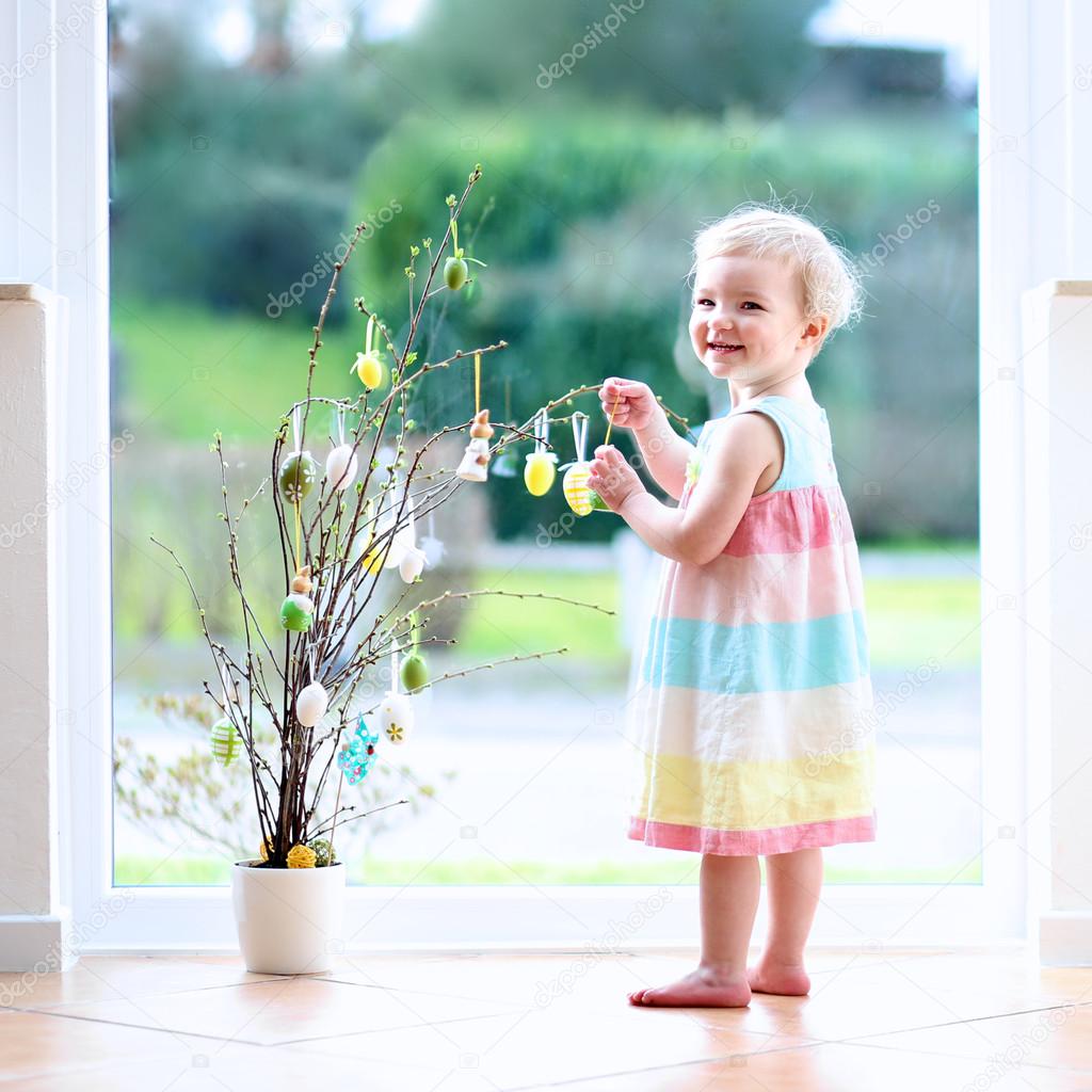 Cheerful blonde little toddler girl decorating cherry tree branches with Easter eggs standing indoors next to a big window with street view