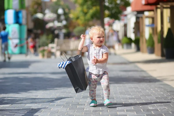 Cute baby girl standing or walking in the middle of the street in outlet village during sales with black shopping bag in her hands, crowd of people in the background — Stock Photo, Image