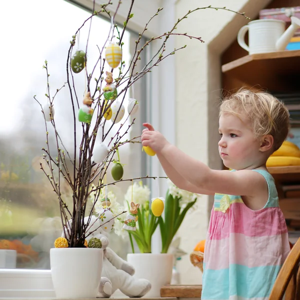 Adorable niña rubia pequeña decoración con huevos de Pascua ramas de cerezo de pie en la cocina al lado de una ventana con vista al jardín — Foto de Stock