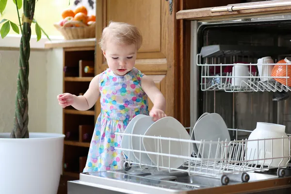 Lachende blonde peuter meisje helpt in de keuken nemen platen uit schotel wasmachine — Stockfoto