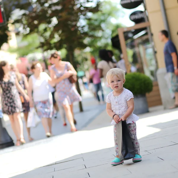 Linda niña de pie o caminando en el medio de la calle en la aldea de salida durante las ventas con bolsa de compras negro en sus manos, multitud de personas en el fondo —  Fotos de Stock