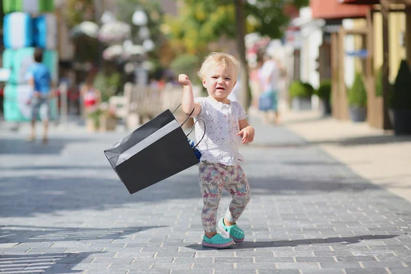 Cute baby girl standing or walking in the middle of the street in outlet village during sales with black shopping bag in her hands, crowd of people in the background — Stock Photo, Image