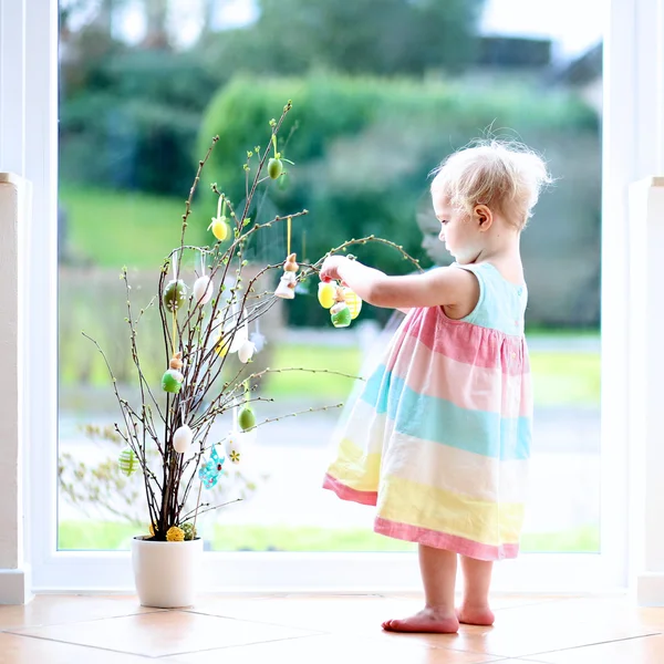 Alegre niña rubia pequeña decoración de ramas de cerezo con huevos de Pascua de pie en el interior junto a una gran ventana con vista a la calle — Foto de Stock